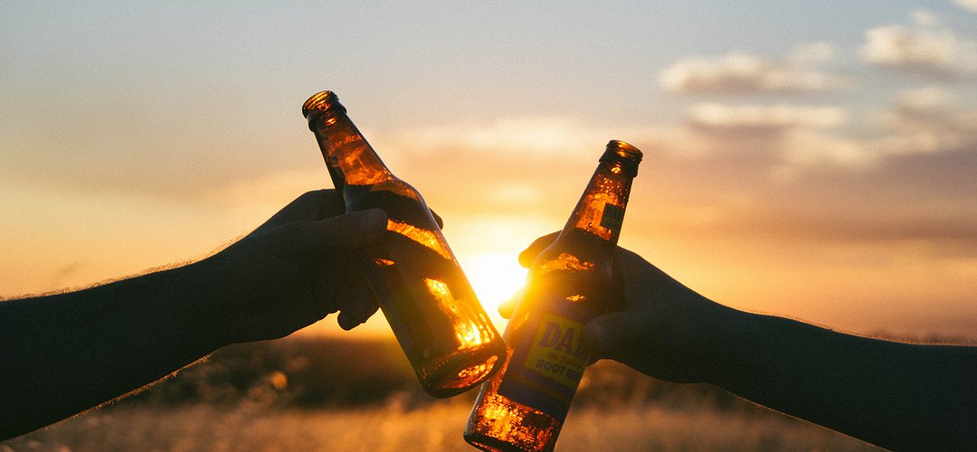 Photographie de deux personnes trinquant avec des bouteilles de bière durant un coucher de soleil.